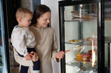 Bir anne, cam vitrindeki çeşitli tatlılara işaret ederken çocuğunu kucağına alır. Bir pastanede sergilenen şekerlerle ilgileniyor gibiler..