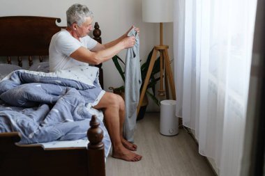 Yaşlı bir adam yatağın kenarında oturur, giysileri katlar ve pencereden süzülen yumuşak sabah ışığının tadını çıkarır..