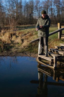 Bir adam ahşap bir platformda duruyor, sakin bir su kütlesinde balık tutuyor. Sabahın erken saatlerinde, sakin manzarayı ve odaklanmış ifadesini vurgular..