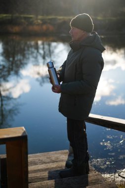 Huzurlu bir gölün yanındaki ahşap rıhtımda bir adam duruyor. Sıkı giyinmiş ve elinde termosla doğanın huzurlu manzarasının tadını çıkarıyor..