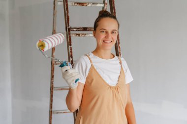 Neşeli bir kadın elinde boya silindiri olan bir merdivenin yanında duruyor, bir ev geliştirme projesi sırasında ışık dolu bir odada boyamaya hazır..