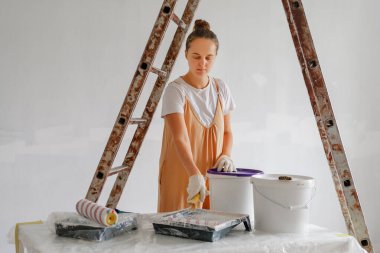 Genç bir kadın, parlak bir odadaki merdivenin yanındaki boya malzemelerini hazırlamaya odaklanmış. Eldiven takıyor ve özenle çalışıyor. Ev geliştirme çabalarını gösteriyor..