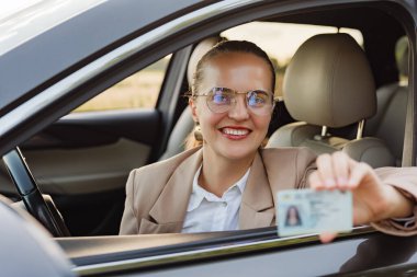 Bir kadın arabada oturuyor, gülümsüyor ve kimlik kartını tutuyor. Güneş parlıyor ve arka planda ağaçlar görülebilir..