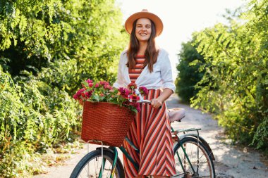 Çevresi yeşilliklerle çevrili bir yolda bisiklet süren bir kadın gülümsüyor. Bisikletinde çiçeklerle dolu bir sepeti var ve şapka takıyor..