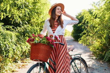 Çevresi yeşilliklerle çevrili bir yolda bisikletinin yanında duran bir kadın. Çiçeklerle dolu bir sepet tutuyor ve gün ışığında gülümsüyor..