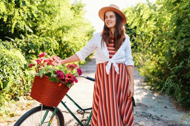 Yolda elinde bir sepet çiçekle bisikletin yanında duran bir kadın. Güneş ışığı yaprakların arasından süzülüp, parlak bir atmosfer yaratır..