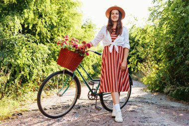 Bir kadın yemyeşil bitkiler arasındaki toprak yolda bisikletinin yanında duruyor. Elinde çiçeklerle dolu bir sepet tutuyor ve kameraya gülümsüyor..