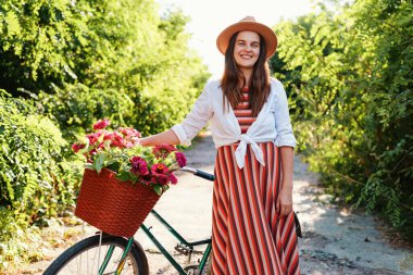 Genç bir kadın, parlak bir yaz gününde yeşil çalılarla çevrili bir yolda elinde bir sepet çiçekle bisiklet tutarken gülümsüyor..