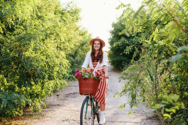 Şapkalı bir kadın yeşilliklerle dolu bir yolda bisiklet sürüyor. Onun bisikletinin çiçeklerle dolu bir sepeti var. Gündüz ve güneşli..