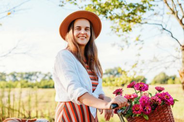 Bir kadın elinde çiçeklerle dolu bir sepet tutan bir bisikletin yanında duruyor. Kırsal alanda güneşli havanın tadını çıkarırken gülümsüyor..