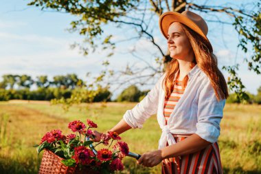 Ağaçlarla ve mavi gökyüzüyle dolu yeşil bir tarlada güneşli bir günün tadını çıkarırken bir kadın elinde çiçeklerle bisiklet sürüyor..