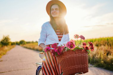 Bir kadın çiçeklerle dolu bir sepetle tarlada bisiklet sürüyor. Güneş arkasından batıyor, parlak bir zemin oluşturuyor..