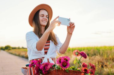 Bir kadın bisikletinin yanında duruyor, sepetteki çiçeklerin fotoğrafını çekerken bir tarlada sıcak bir akşamın tadını çıkarıyor. Gülümserken etrafında parlak çiçekler uçuşuyor..