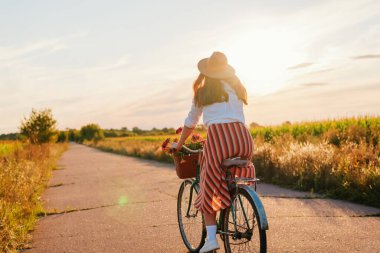 Güneş batarken, elinde çiçeklerle dolu bir sepet tutan bir kadın tarlalarla çevrili sessiz bir yolda bisiklet sürüyor. Gökyüzü ılık renklerle aydınlık..