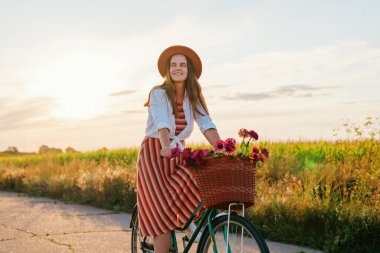 Bir kadın çiçeklerle dolu bir sepetle bisiklete biniyor. Yeşil tarlaları olan kırsal bir alanda günbatımının tadını çıkarırken gülümsüyor..