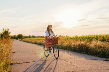 Kırsal bir yolda elinde çiçekli bir sepetle bisiklet süren bir kadın. Güneş ufukta batarken, sıcak havanın tadını çıkarıyor..