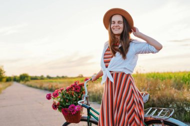 Gün batımında kırsalda bir yolda elinde bir sepet çiçekle bisikletin yanında duran bir kadın. Anın tadını çıkarırken gülümsüyor..