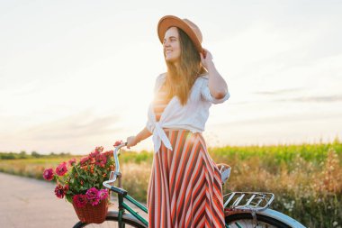 Kadın saçlarını tutarken ve bisikletinin yanında dikilirken sepetteki çiçeklerle gülümsüyor. Ekinlerle dolu büyük bir tarlanın arkasında güneş batar..