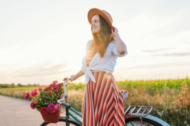 Bir kadın şapkasını düzeltirken gülümsüyor, sepette çiçeklerle bir bisikletin yanında duruyor. Güneş arkasından batıyor, tarlaya sıcak bir parıltı saçıyor..