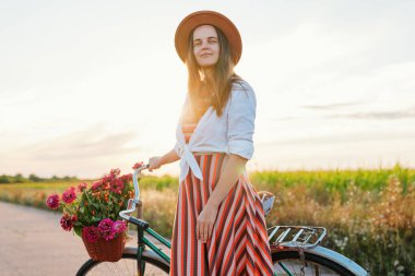 Kırsal bir yolda, gün batımında yeşil tarlalar ve açık bir gökyüzü ile çevrili, sepetinde çiçek açan bir kadın bisikletinin yanında duruyor..