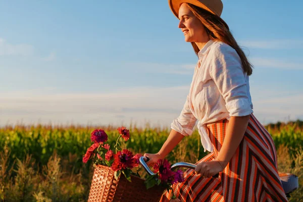 Bir kadın açık arazide bisiklet sürüyor. Sepetinde çiçekler var ve güneş arka planda batıyor. Sıcak bir ışık yaratıyor..