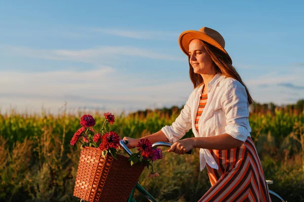 Genç bir kadın bir sepet dolusu çiçekle bisiklet sürüyor. Bir tarlanın yakınındaki yolda bisiklet sürerken sıcak akşam ışığından zevk alır..