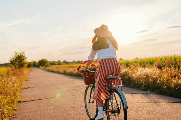 Güneş batarken, elinde çiçeklerle dolu bir sepet tutan bir kadın tarlalarla çevrili sessiz bir yolda bisiklet sürüyor. Gökyüzü ılık renklerle aydınlık..
