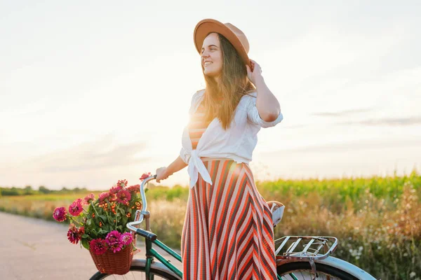 Kadın saçlarını tutarken ve bisikletinin yanında dikilirken sepetteki çiçeklerle gülümsüyor. Ekinlerle dolu büyük bir tarlanın arkasında güneş batar..