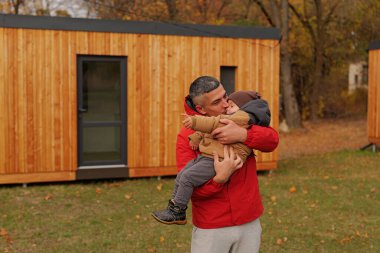 Bir adam çocuğunu renkli sonbahar yaprakları olan açık bir alanda sevgiyle kucağına alır. Arkadaki ahşap kulübeler rahat bir atmosfer yaratıyor..