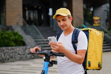 Teslimat işçisi mobiletin üzerinde duruyor ve şapka takıp sırt çantası taşıyan bir telefona bakıyor. Mekan kalabalık bir kentsel alan..
