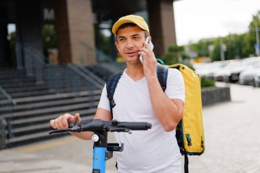 Adam elektrikli scooter 'ın yanında duruyor, kulağına telefon dayayıp şehir ortamında sarı bir şapka ve sırt çantası takıyor..