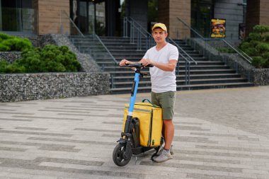 Teslimat işçisi alışveriş merkezinin dışında elektrikli scooterın üzerinde duruyor. Günlük kıyafetler giyiyor ve motosikletine içinde yiyecek olan sarı bir çantası var..