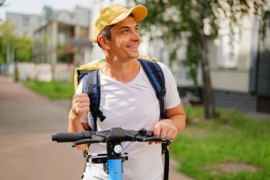 Adam, güneşli bir günde şehrin bir bölgesine yemek götürmeye hazırlanırken sırt çantasıyla elektrikli scooter 'a biner ve gülümser..