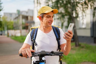Şehrin caddesinde elektrikli scooter kullanan bir adam telefonuna bakıyor. Şapka takıyor ve sırt çantası var. Arkada ağaçlar ve binalar var..
