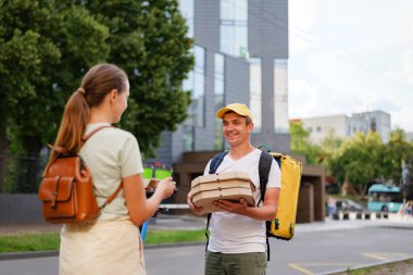 Teslimatçı, güneşli bir günde bir sırt çantası taşıyarak modern bir binanın dışındaki müşteriye paketlenmiş yiyecek verir..