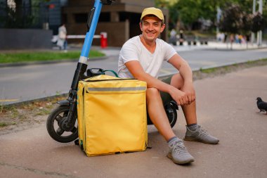 Teslimat işçisi, şehrin yoğun bir bölgesinde kaldırımda elektrikli bir scooter ve yiyecek teslimatı için sarı bir çantanın yanında oturuyor..