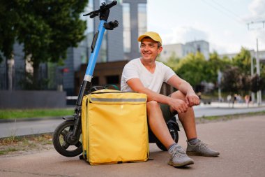 Teslimatçı, öğleden sonra elinde bir teslimat çantasıyla bir elektrikli scooter 'ın yanında dinleniyor. Şehir ortamının arka planında binalar var.