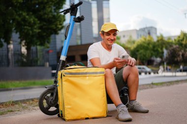 Bir teslimatçı şehir parkında yerde oturuyor. Telefonunu motosiklet ve büyük sarı bir teslimat çantasının yanında dinlenirken kullanıyor..