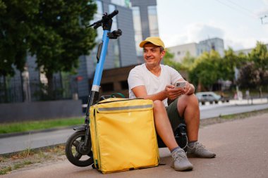 Teslimat işçisi, şehir caddesinde motosiklet ve sarı termal çantanın yanında otururken telefonu kontrol ediyor.