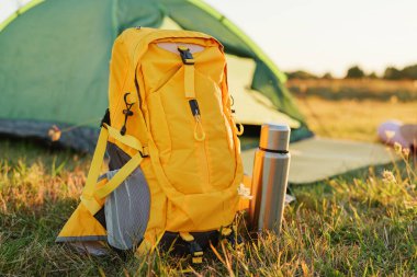 Sarı bir sırt çantası bir kamp çadırının yanındaki çimlerin üzerinde dinleniyor. Arka planda güneş batarken yakınlarda gümüş bir su şişesi duruyor..