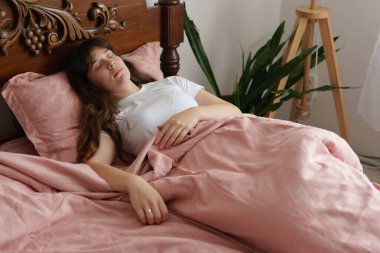 Uzun saçlı bir kadın, yumuşak pembe battaniyeler ve parlak, davetkar bir uzayda yapraklı bir bitki ile çevrili, rahat bir yatakta huzur içinde uyur..