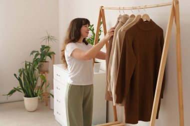 Basit beyaz gömlekli ve yeşil pantolonlu bir kadın, parlak bir odada, parlak bitkiler ve açık renk dekorlu ahşap bir askıda rahat süveterler düzenler..