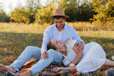 İki kişi bir tarlada battaniyenin üzerinde rahatlar. Biri yanında yatarken diğeri oturur. Gülümsemeleri paylaşıyorlar ve doğal bir ortamda birbirlerine bağlanıyorlar..