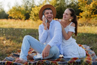 Bir adam ve bir kadın, güneşli bir günde ağaçların altındaki çimlerin altında bir battaniyede oturup gülümsüyor ve telefon görüşmesi yapıyorlar..