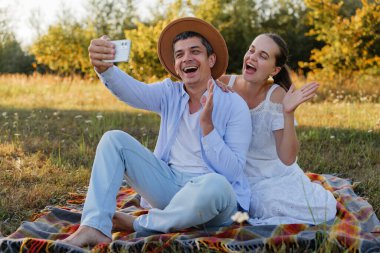 İki kişi tarlada bir battaniyeye oturup gülümsüyor ve selfie çekiyor. Güneşli bir öğleden sonra sıcak havadan ve birbirlerinin arkadaşlığından hoşlanırlar..