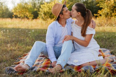 İki insan çimenli bir tarlada battaniyenin üzerinde oturmuş, gecenin sıcak güneşi altında öpüşürken birbirlerinin varlığının tadını çıkarıyorlar..