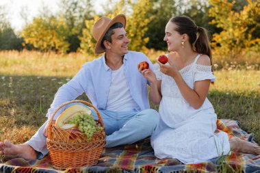 Erkek ve kadın bir tarlada battaniyeye oturur. Elma tutarken birbirlerine gülümserler. Çimlerin üzerinde bir sepet meyve var..