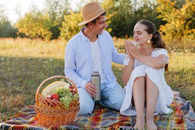 Parkta bir çift battaniyeye oturur. Meyve dolu bir sepetteki yiyecekleri ve termostan içecekleri paylaşıyorlar. Çevrede yeşil yapraklı ağaçlar var..