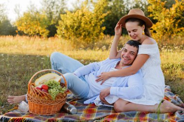 Bir çift tarlada bir battaniyeye oturmuş, sepetteki meyveleri paylaşırken gülümsüyor ve gülüyor. Güneş doğadaki mutlu anlarında parlıyor..