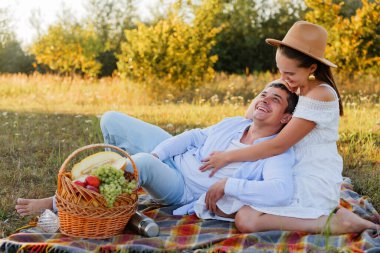 İki kişi çim ve ağaçlarla dolu bir tarlada bir battaniyede oturuyor. Meyvelerle piknik yaparlar ve güneşin altında birlikte gülerler..